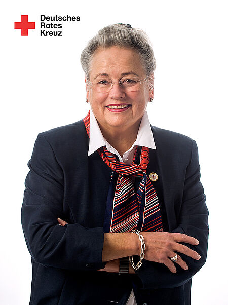 Person mit verschränkten Armen trägt dunkles Sakko, weißes Hemd und gestreiften Schal vor weißem Hintergrund mit Logo des Deutschen Roten Kreuzes