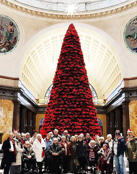 Große Gruppe von Menschen vor einem hohen, rot geschmückten Weihnachtsbaum in einer prunkvollen Halle mit Kuppeldecke.